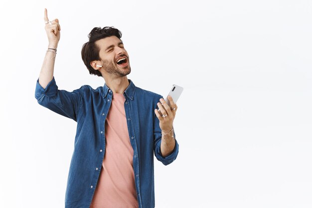 ragazzo con gli auricolari wireless che ascolta musica, alza la mano cantando lungo i testi delle canzoni preferite, tenendo in mano lo smartphone, goditi la straordinaria qualità delle cuffie