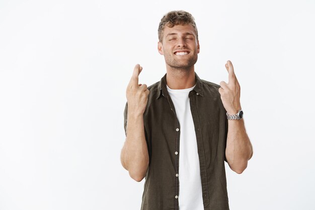 ragazzo con i capelli biondi e il sorriso bianco che sorride felice con gli occhi chiusi mentre esprime un desiderio con le dita incrociate per buona fortuna, si spera pregando.