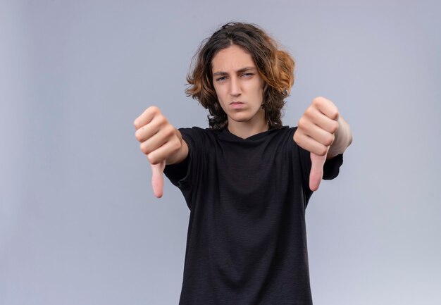 Ragazzo con i capelli lunghi in maglietta nera mostra il pollice verso il basso sul muro bianco