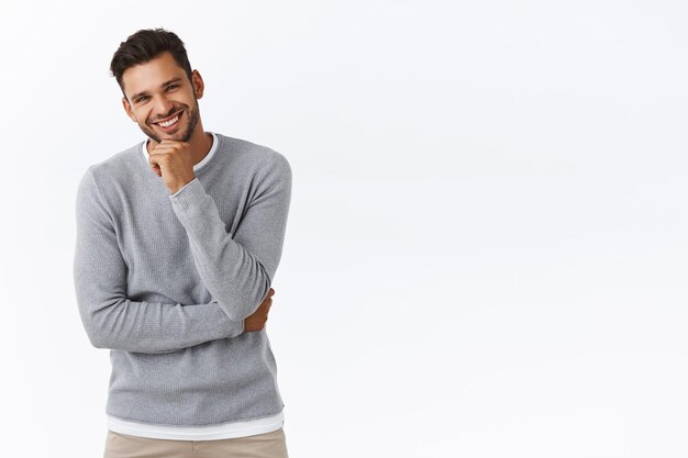 ragazzo con la barba in maglione grigio sopra la maglietta, ridendo, sorridendo felicemente, discutendo di un momento interessante della vita, strofinando il mento e guardando interessato, riflettendo sull'idea