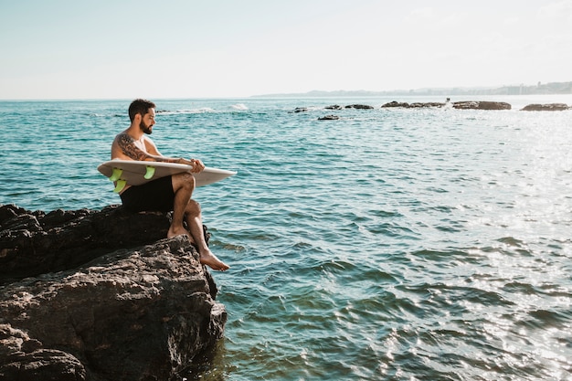 Ragazzo con tavola da surf che si siede sulla roccia vicino al mare