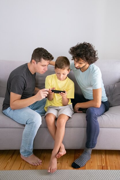 Ragazzo concentrato che gioca gioco sul telefono cellulare, i suoi due papà seduti vicino a lui e aiutando. Colpo verticale. Famiglia a casa e concetto di comunicazione