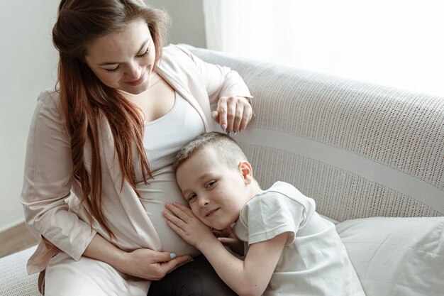 Ragazzo del bambino che abbraccia la pancia della gravidanza di sua madre sul divano di casa.