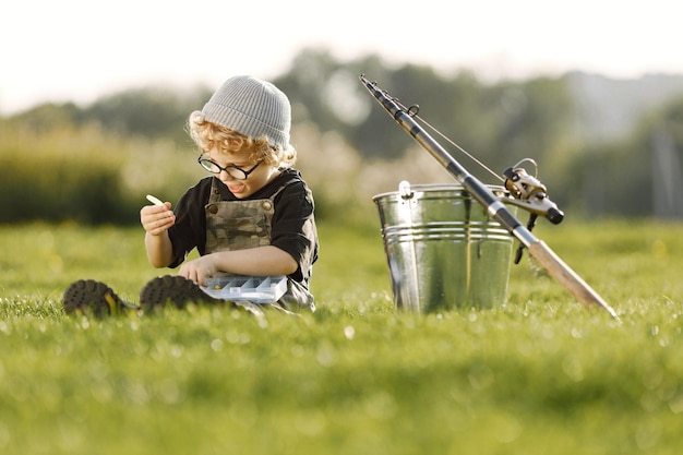 Ragazzo del bambino che tiene una scatola con un'esca per la pesca Ragazzo che indossa tuta color cachi Ragazzo seduto su un'erba vicino al secchio e alla canna da pesca