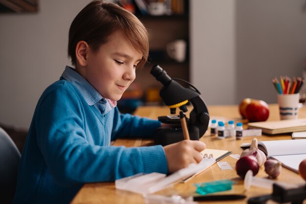 Ragazzo del primo grado che studia a casa usando il microscopio