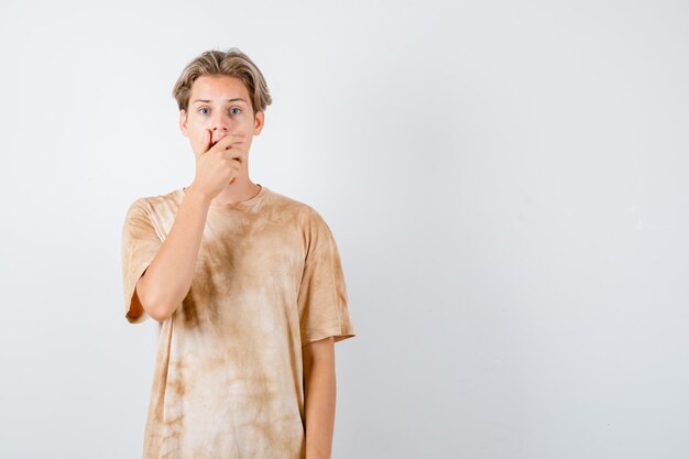 Ragazzo dell'adolescente in maglietta che tiene la mano sulla bocca e sembra deluso, vista frontale.