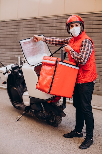 Ragazzo delle consegne di cibo alla guida di uno scooter con scatola con cibo e maschera da indossare