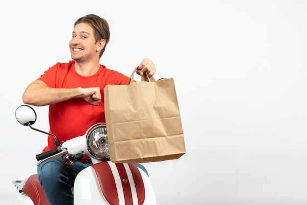 Ragazzo di corriere felice sorridente dei giovani in uniforme rossa che si siede sul sacchetto di carta della tenuta del motorino sulla parete bianca