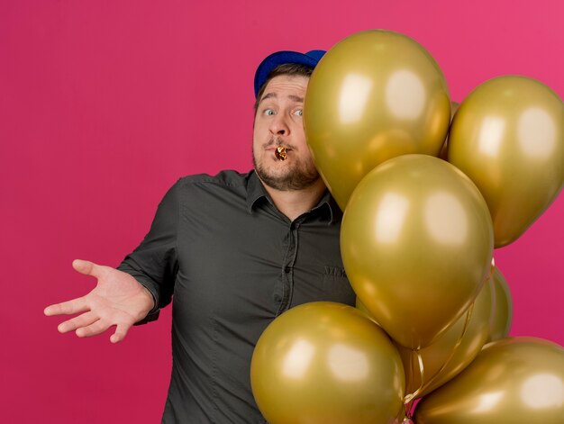 Ragazzo di partito giovane confuso che indossa cappello blu in piedi vicino a palloncini che tengono il ventilatore del partito in bocca che diffonde le mani isolate sul colore rosa