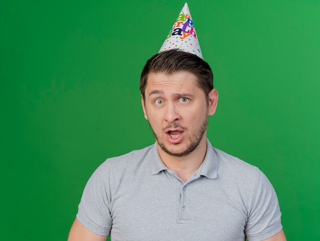 Ragazzo di partito giovane confuso che indossa il cappello di compleanno isolato sul verde