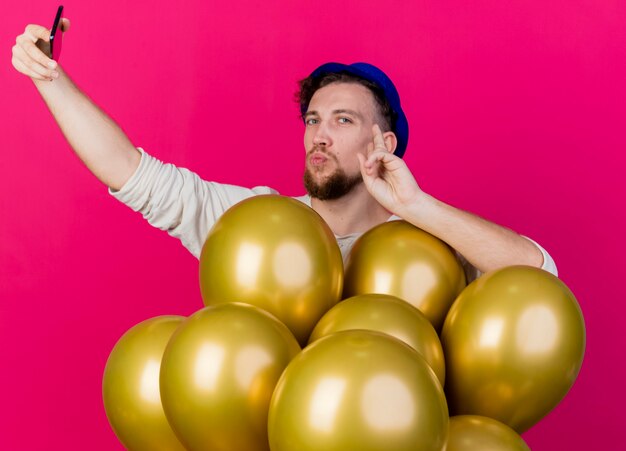 Ragazzo di partito slavo bello che indossa il cappello del partito in piedi dietro i palloncini che guarda l'obbiettivo che tocca il fronte prendendo selfie isolato su priorità bassa cremisi