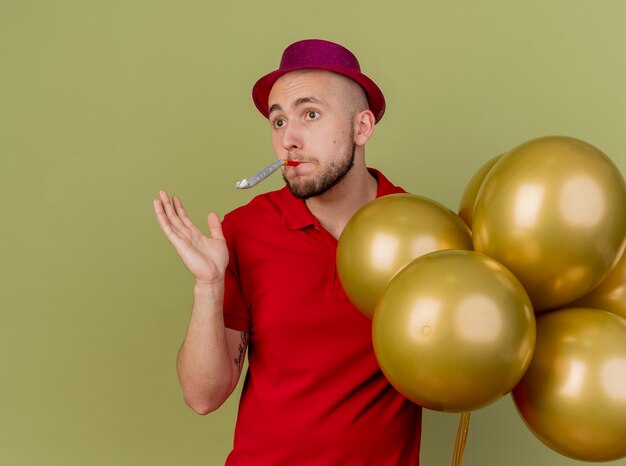 Ragazzo di partito slavo bello colpito giovane che indossa il cappello del partito che tiene palloncini che soffia il ventilatore del partito che mostra la mano vuota che esamina il lato isolato su fondo verde oliva con lo spazio della copia