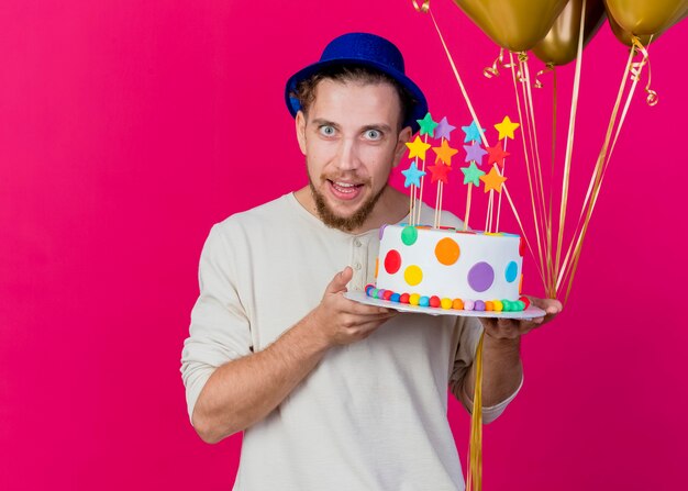 Ragazzo di partito slavo bello colpito giovane che indossa il cappello del partito che tiene palloncini e torta di compleanno con le stelle che guarda l'obbiettivo isolato su priorità bassa cremisi con lo spazio della copia