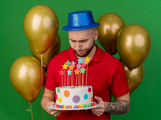 Ragazzo di partito slavo bello giovane dispiaciuto che indossa il cappello del partito in piedi davanti a palloncini che tengono e guardando la torta di compleanno isolata su priorità bassa verde