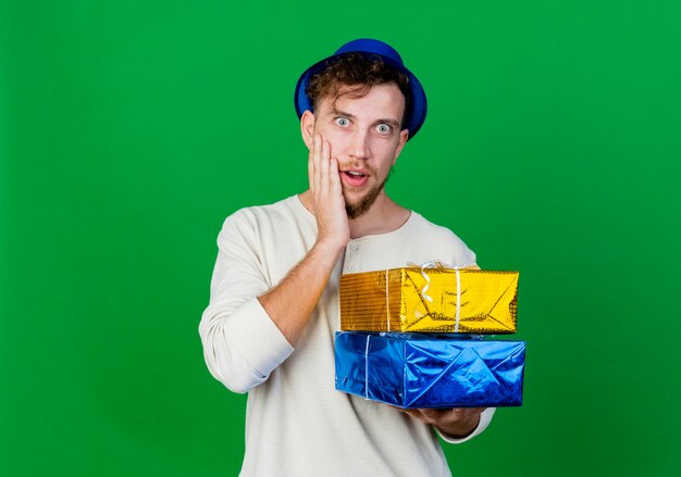 Ragazzo di partito slavo bello giovane sorpreso che indossa il cappello del partito che tiene i contenitori di regalo che guarda l'obbiettivo che tiene la mano sulla faccia isolata su fondo verde con lo spazio della copia