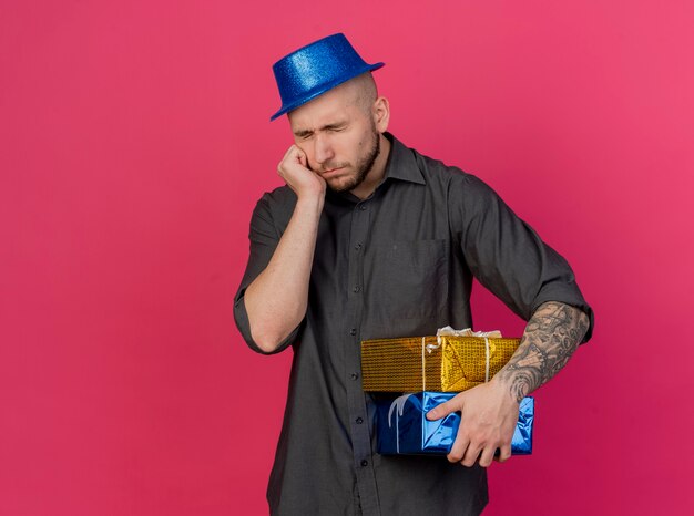 Ragazzo di partito slavo bello giovane triste che indossa il cappello del partito che tiene confezioni regalo e sacchetti di carta isolati su sfondo cremisi con spazio di copia