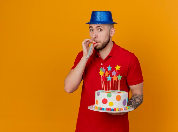 Ragazzo di partito slavo bello impressionato che indossa il cappello del partito che guarda l'obbiettivo che tiene la torta di compleanno con il ventilatore del partito in bocca isolato su priorità bassa arancione con lo spazio della copia