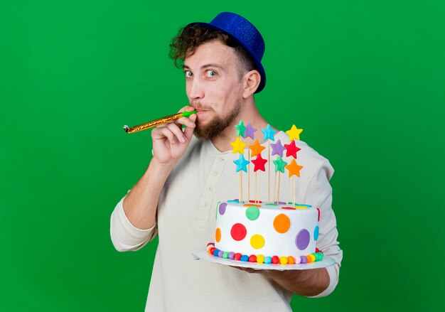 Ragazzo di partito slavo bello impressionato che indossa il cappello del partito che tiene la torta di compleanno con le stelle che soffia il ventilatore del partito che guarda l'obbiettivo isolato su priorità bassa verde con lo spazio della copia