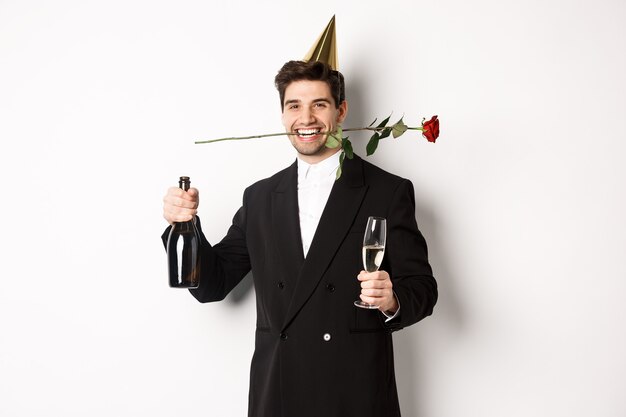 Ragazzo divertente in abito alla moda, celebrando e facendo una festa, tenendo rosa tra i denti e champagne, in piedi su sfondo bianco.
