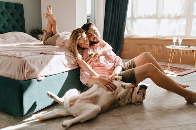 Ragazzo e la sua ragazza stanno riposando in camera da letto. Coppie felici che esaminano amorevolmente il loro animale domestico che vuole giocare.