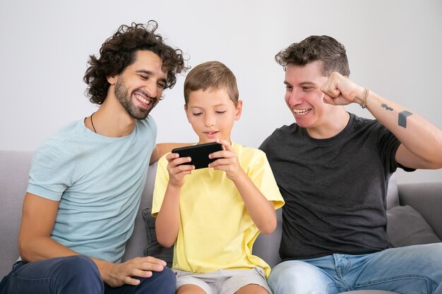 Ragazzo eccitato che gioca sul cellulare, i suoi due papà felici seduti vicino a lui e aiutando. Vista frontale. Famiglia a casa e concetto di comunicazione