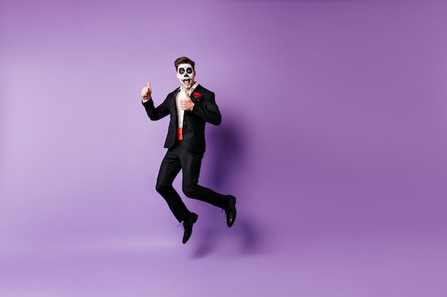 Ragazzo eccitato e ben vestito con trucco spaventoso che scherza in studio Modello zombie divertente che salta su sfondo viola