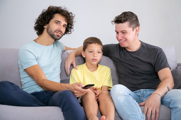 Ragazzo eccitato e i suoi allegri papà che guardano programmi TV a casa, seduti sul divano in soggiorno, abbracciati, usando il telecomando, distogliendo lo sguardo. Famiglia e concetto di intrattenimento domestico