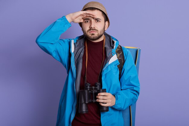 Ragazzo escursionista turistico con zaino e tappetino da viaggio, indossa giacca e cappello, sta con il binocolo sul collo