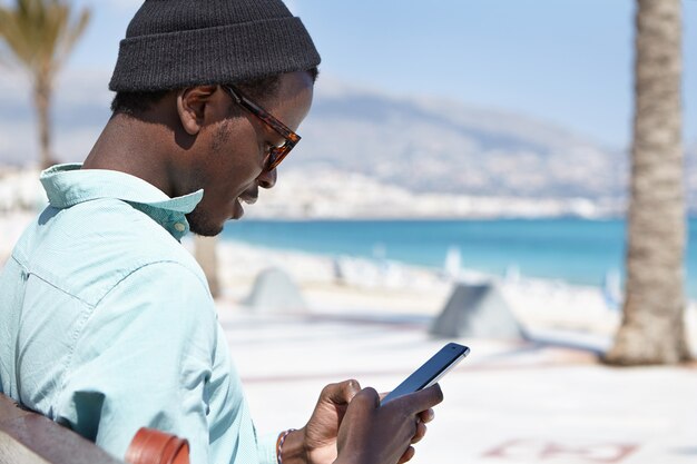 Ragazzo europeo nero alla moda attraente che si distende durante il giorno, seduto su una panchina vicino al mare, tenendo e utilizzando un moderno dispositivo elettronico per il networking, godendo la comunicazione online con gli amici