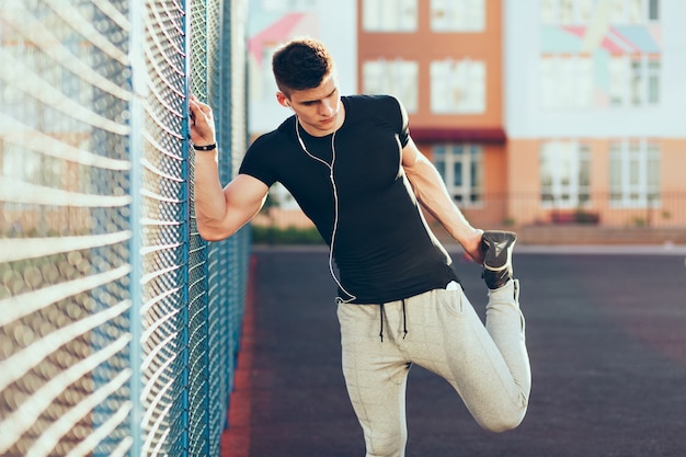 Ragazzo forte con un corpo muscoloso in allenamento vicino al recinto al mattino sullo stadio. Indossa abiti sportivi, ascolta la musica con le cuffie.