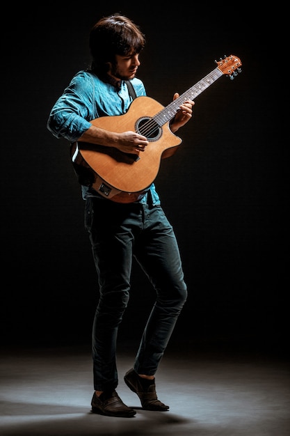 Ragazzo freddo con il cappello in piedi con la chitarra su sfondo scuro studio