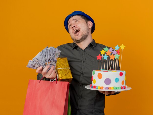 ragazzo gioioso partito giovane che indossa il cappello blu tenendo la torta con doni e denaro isolato su arancione