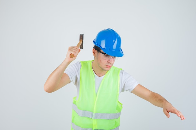 Ragazzo giovane azienda martello in costruzione uniforme e guardando concentrato. vista frontale.