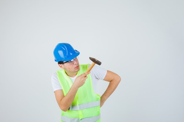 Ragazzo giovane azienda martello, mettendo la mano sulla vita, guardando il martello in uniforme da costruzione e guardando concentrato. vista frontale.