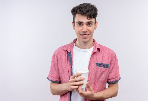Ragazzo giovane caucasico sorridente che porta la camicia rosa che tiene tazza di caffè su fondo bianco isolato