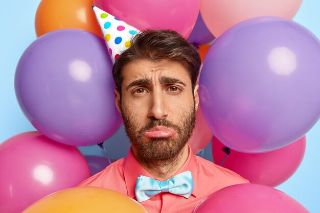 Ragazzo giovane infelice in posa circondato da palloncini colorati compleanno