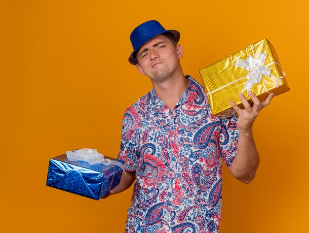 Ragazzo giovane partito dispiaciuto che indossa il cappello blu che tiene i contenitori di regalo isolati sull'arancio