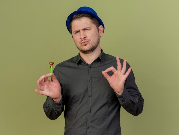 Ragazzo giovane partito impressionato che indossa camicia nera e cappello blu che tiene il ventilatore del partito che mostra il gesto okey isolato su verde oliva