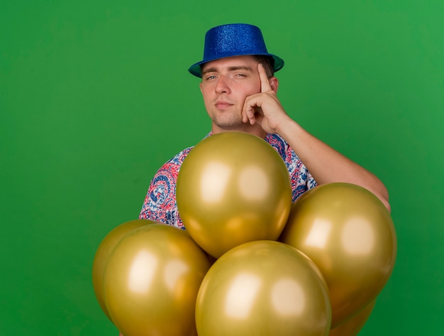 Ragazzo giovane partito impressionato che indossa cappello blu in piedi dietro palloncini e mettendo la mano sul tempio isolato sul verde