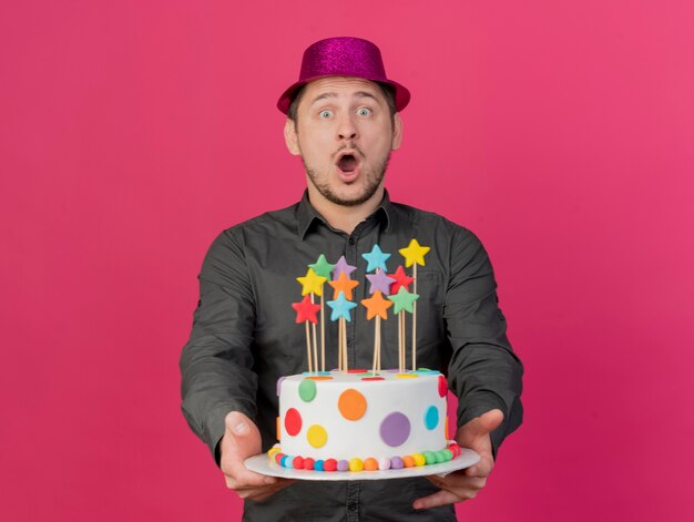 Ragazzo giovane partito sorpreso che porta il cappello dentellare che tiene fuori la torta isolata sul colore rosa