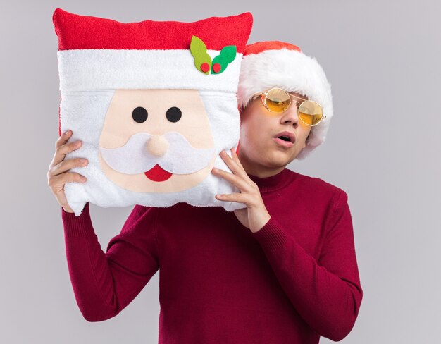 Ragazzo giovane sorpreso che indossa il cappello di Natale con gli occhiali che tengono il cuscino di Natale isolato su priorità bassa bianca