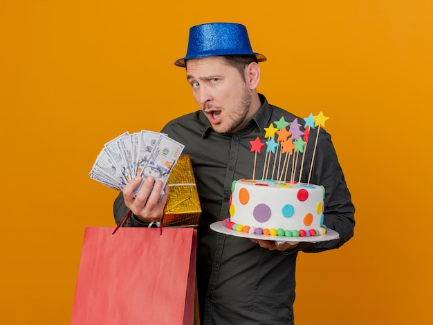Ragazzo giovane sorpreso del partito che porta i regali blu della tenuta del cappello con la torta ed i contanti isolati sull'arancia