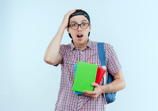 Ragazzo giovane sorpreso dell'allievo che indossa la borsa posteriore e gli occhiali e il berretto che tengono i libri e che mette la mano sulla testa su bianco