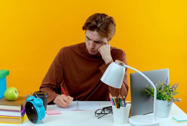 Ragazzo giovane studente di pensiero che si siede allo scrittorio con gli strumenti della scuola che scrivono qualcosa sul taccuino su colore giallo