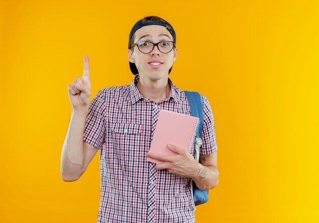 Ragazzo giovane studente impressionato che indossa zaino e occhiali e berretto che tiene taccuino e punta verso l'alto