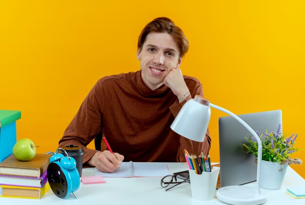 Ragazzo giovane studente sorridente che si siede allo scrittorio con gli strumenti della scuola che scrivono qualcosa sul taccuino su colore giallo