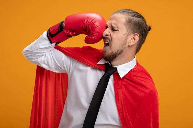 Ragazzo giovane supereroe che indossa cravatta e guantoni da boxe che si batte isolato su sfondo arancione