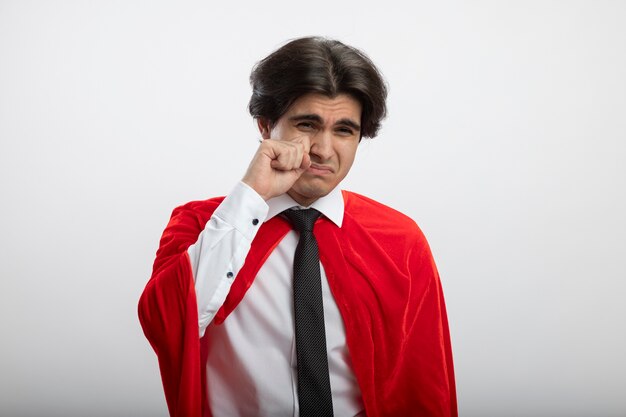 Ragazzo giovane supereroe dispiaciuto che guarda l'obbiettivo che indossa cravatta asciugandosi gli occhi con la mano isolata su bianco