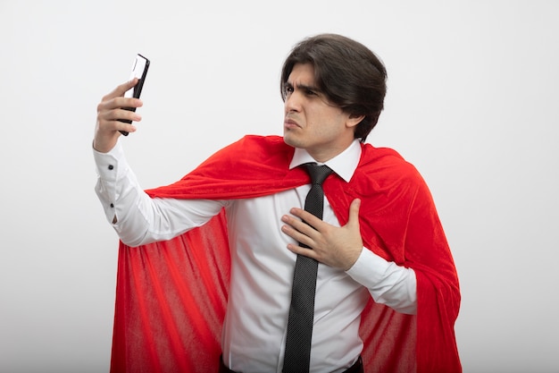 Ragazzo giovane supereroe dispiaciuto che indossa cravatta holding e guardando il telefono mettendo la mano sul petto isolato su sfondo bianco