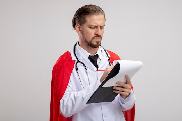 Ragazzo giovane supereroe rigoroso che indossa abito medico con lo stetoscopio scrivendo qualcosa negli appunti isolato su bianco